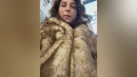 Video of a woman with fair skin and shoulder-length brown hair, wearing a brown fur coat, standing in a brightly lit room with a window and abstract art on the wall.