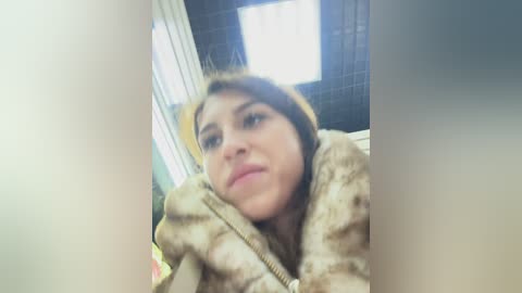 Video of a young woman with light skin and shoulder-length brown hair, wearing a brown and beige fur coat, looking introspective in a dimly lit room with tiled walls and a window.