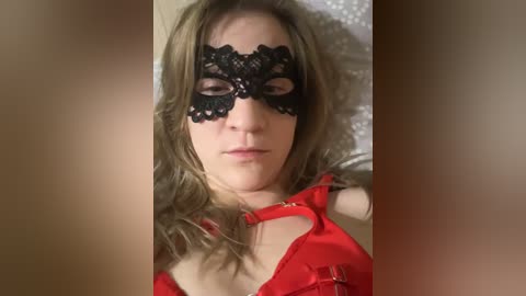 Video of a fair-skinned woman with long, wavy blonde hair, wearing a black lace masquerade mask and a red satin dress. She lies on a white bed with a blurred background.