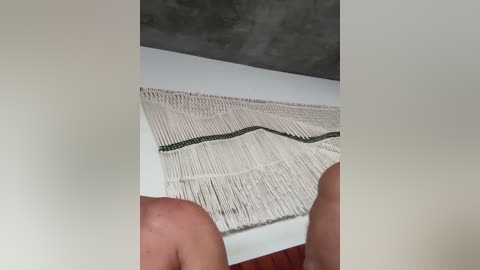 Video of a person's hands weaving a beige, fringed tapestry with a black and green pattern on a white surface, set against a grey concrete wall.