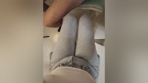A video captures a person wearing light gray jeans and a white shirt, seated on a green chair. The background shows a tiled floor and part of a white cabinet.