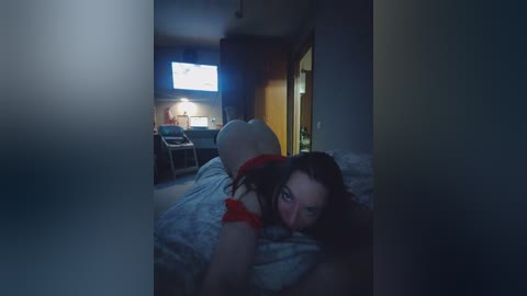 Video of a young woman with dark hair, lying on a bed in a dimly lit bedroom. She wears a red top and has her back exposed. A TV screen is visible in the background.