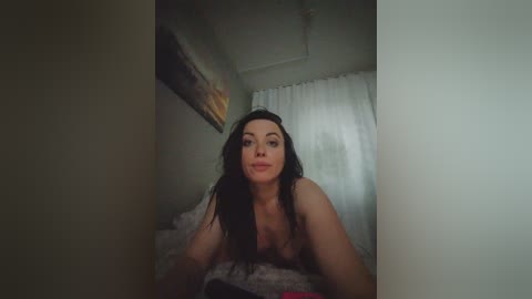 Video of a nude woman with long black hair, light skin, and medium breasts, lying on a bed with white curtains in the background, in a dimly lit room.