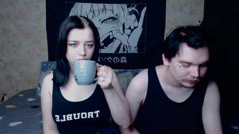 Video of two young people, a woman with dark hair, wearing a black \"TORA\" shirt, and a man with short hair, wearing a black tank top, sitting on a bed, both holding mugs, in a dimly lit room with a poster of a manga character on the wall.