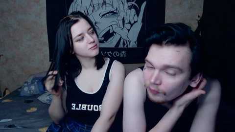 Video of a young woman with long black hair in a black tank top, leaning against a man with short dark hair and a beard, both resting on a bed with Japanese anime posters on the wall.