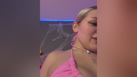 A video of a light-skinned woman with blonde hair in a pink, sleeveless top, wearing a silver necklace. Background features a spider web decoration and a pink and purple light strip.