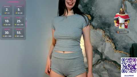 Video of a smiling woman with fair skin and long black hair, wearing a light gray crop top and matching shorts, standing in front of a textured wall with a QR code in the bottom right.