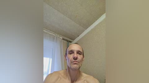 A video of a bald, muscular, shirtless man with a light beard and serious expression standing in a beige room with textured ceiling and white curtains.
