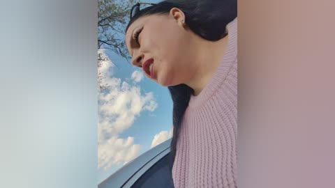 A video of a young woman with long black hair, wearing a pink knitted sweater, looking out a car window at a bright blue sky with scattered white clouds.