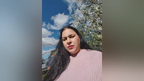 Video of a plus-size woman with long black hair, wearing a pink knit top, standing outdoors under a clear blue sky with fluffy white clouds and blooming trees in the background.