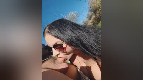 A video of a topless woman with long black hair, wearing sunglasses, kissing a shirtless man. They are outdoors under a clear blue sky, surrounded by autumn foliage.