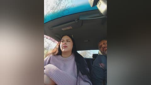 A video of a young woman in a lavender sweater sitting in a car, looking contemplative, with a man in the background smiling.