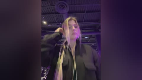 A video of a blonde woman in a black jacket adjusting her hair, standing in an industrial-looking room with exposed pipes and a metallic ceiling.