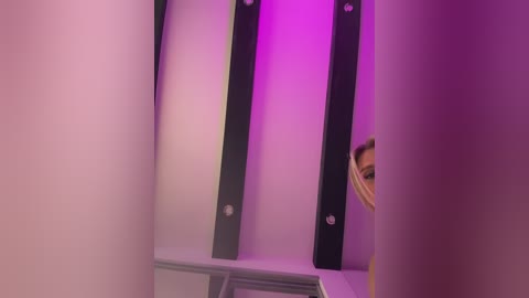 Video of a minimalist interior with a black metal shelf mounted against a white wall, illuminated by purple lighting, partially obscured by a beige curtain.