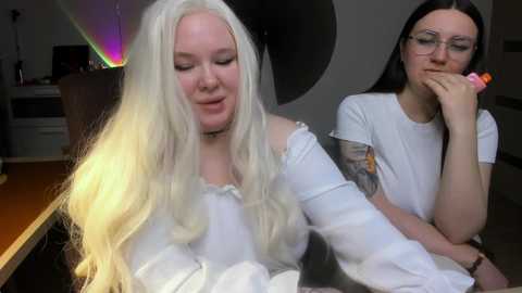 A video shows two women with long hair in a dimly lit room; one wears glasses and the other has a tattoo, both in casual attire.
