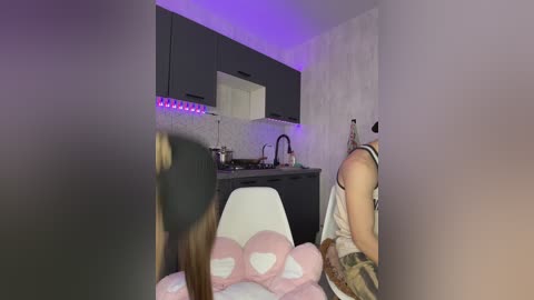 Video of a modern kitchen with purple LED lighting, black cabinets, and a white tiled backsplash. A person in a black cap and plaid shirt is seated at a white chair, holding pink plush toys.