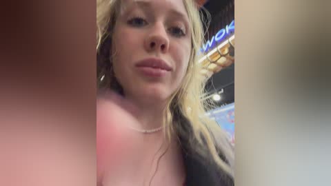 Video of a blonde woman with fair skin, wearing a pink top, smiling slightly, indoors with a blurred background featuring a neon sign.