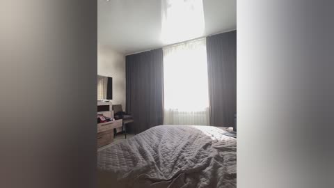 Video of a minimalist, modern bedroom with a large window draped in dark gray curtains, a white bed with a gray quilt, a wooden dresser, and a black chair. The room is bathed in soft natural light.