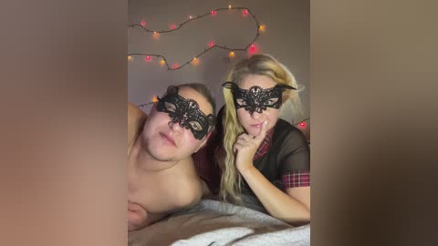 Video of a topless man and a woman with a black lace mask and long blonde hair, lying on a bed with a festive string of lights in the background.