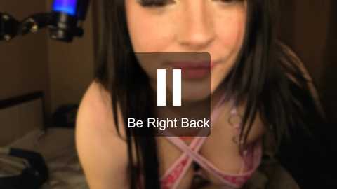 Video of a young woman with long black hair and fair skin, wearing a pink bra, kneeling on a bed with a dimly lit room in the background. Text overlay: \"Be Right Back.\