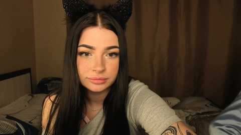 Video of a young woman with long black hair, wearing black cat ear headbands and a grey top, seated on a bed with a beige curtain backdrop.