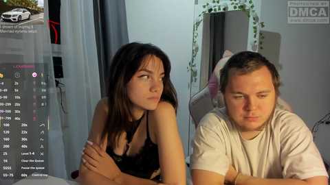 Video of a brunette woman in black lingerie and a bearded man in a white t-shirt, sitting close, indoors, with a TV screen showing a live stream in the background.