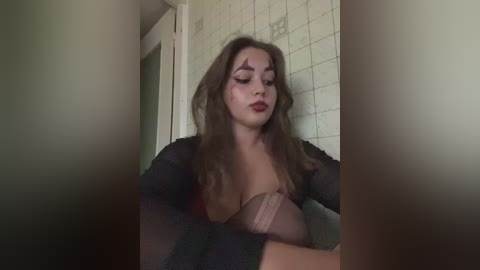 Video of a young woman with long brown hair, wearing a sheer black robe, sitting on a tiled bathroom floor. She has dark makeup and a large, tan-colored prosthetic breast.