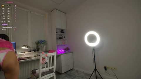 Video of a woman in a pink bikini, sitting at a white desk, illuminated by a ring light, in a minimalist white room with a closed window.
