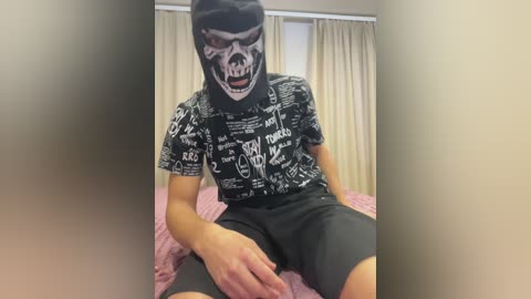 Video of a person wearing a black mask with a skull design, a black T-shirt with white text, and gray shorts, sitting on a bed with pink sheets, in a room with beige curtains.