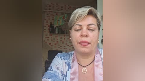 Video of a middle-aged woman with short, light brown hair and fair skin, wearing a light blue tie-dye shirt and a pink necklace, smiling with closed eyes in a cozy, brick-wall room.