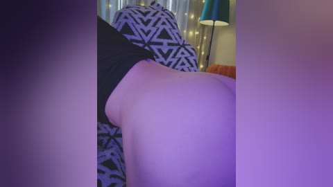 Video of a close-up of a person's buttocks, fair skin, wearing black shorts, lying on a bed with geometric-patterned sheets and a teal lamp in the background.