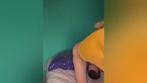 Video of a blonde woman in a yellow shirt and purple pajamas lying on a bed with a teal wall in the background.