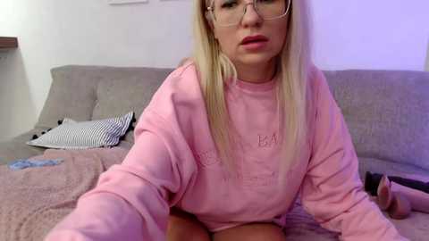 A video of a blonde woman with glasses, wearing a pink hoodie, lying on a grey couch in a dimly lit room.