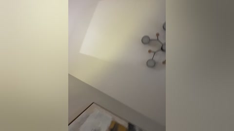 Video of a modern, minimalist interior featuring a white ceiling with a pendant light fixture. The light casts soft shadows, creating a serene atmosphere. A small, abstract artwork with geometric shapes is partially visible on the wall.