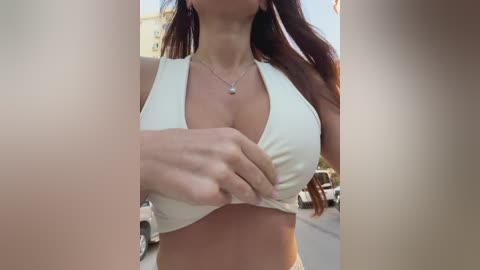 Video of a woman in a white halter top, revealing cleavage, with a silver necklace, standing outdoors near parked cars on a sunny day.