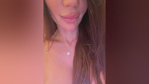 A close-up video of a fair-skinned woman with long, brown hair, wearing a silver necklace with a small, rectangular pendant. The background is a gradient of warm, reddish-brown tones.