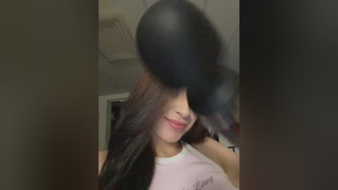 Video of a young Asian woman with long black hair, partially obscured by a cloud of smoke. She has fair skin and wears a white tank top with a small text logo. Background shows a dimly lit room with a door.