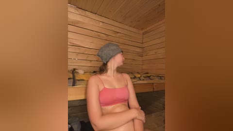 Video of a fair-skinned woman with brown hair, wearing a gray beanie and pink sports bra, sitting in a wooden sauna with wooden benches and towels in the background.