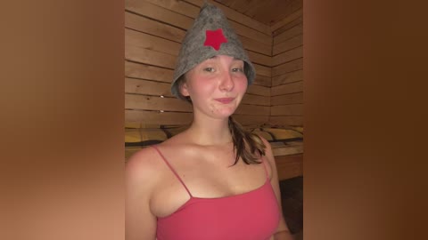 Video of a fair-skinned woman with a slight blush, wearing a grey knit beanie with a red star and a pink spaghetti-strap top, inside a wooden sauna.