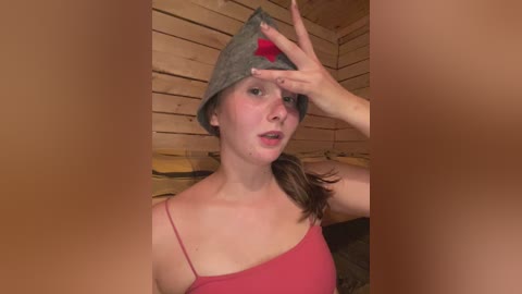 Video of a fair-skinned woman with brown hair, wearing a gray beanie with a red star, and a red tank top, in a wooden cabin with a blurred background.