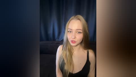 Video of a young Caucasian woman with long blonde hair, wearing a black sleeveless top, standing against dark blue curtains in a dimly lit room.