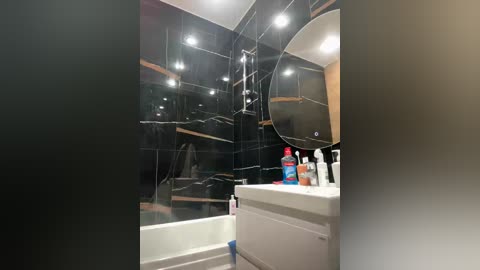 Video of a modern bathroom with black tiled walls, a white bathtub, a white sink with toiletries, and a mirror reflecting the space, under bright ceiling lights.