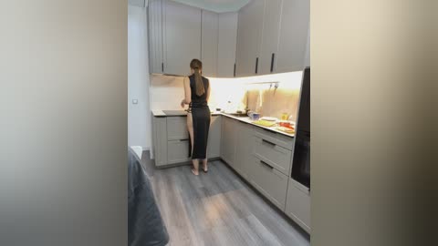 Video of a modern kitchen with minimalist design. A woman in a black dress stands facing the sink, barefoot on wooden floors. Light gray cabinets, stainless steel appliances, and a sleek backsplash are visible.