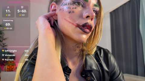 Video of a young woman with fair skin and long blonde hair, wearing a black leather jacket, heavy black makeup, and face paint, in a dimly lit room.