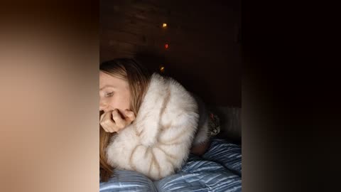 Video of a young woman with long, light brown hair, wrapped in a white, fluffy blanket, lying on a blue and white striped bed. Warm, dimly lit room with a string of fairy lights on the wall in the background.