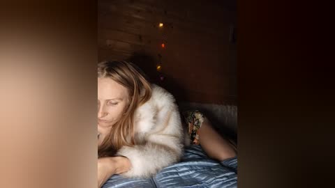 Video of a young Caucasian woman with long brown hair, lying on a bed, wearing a white fur coat, in a dimly lit room with exposed brick walls and string lights.