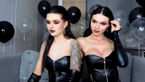 Video of two women with long black hair, wearing black leather corsets, tattoos, and dark makeup, in a modern living room with gray sofa, black balloons, and translucent balloons.