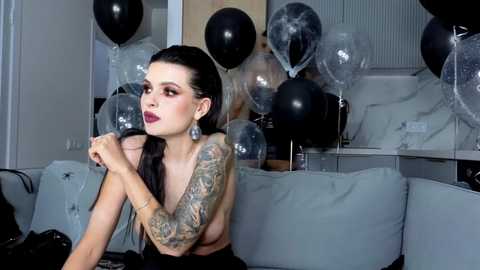 Video of a tattooed, pale-skinned woman with dark hair and dramatic makeup, holding a black balloon, sitting on a grey couch amidst a room filled with black and silver balloons.