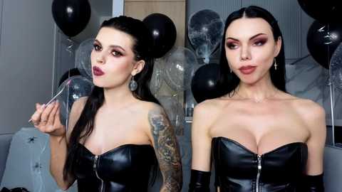 Video of two women with fair skin, long black hair, and dark makeup, wearing black leather corsets, tattoos, and holding black balloons, in a modern, dimly lit room.