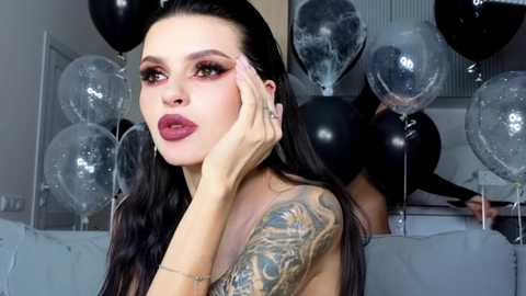 Video of a fair-skinned woman with long black hair, wearing dark lipstick and dramatic makeup, holding a black balloon. She has a detailed tattoo sleeve and is indoors surrounded by other black and clear balloons.